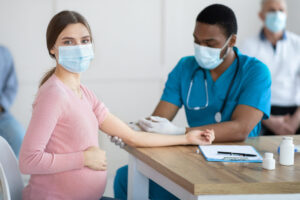 Pregnant woman in face mask getting vaccinated against Covid-19 at clinic. African American doctor giving coronavirus vaccine injection to expectant lady. Healthcare and global immunization