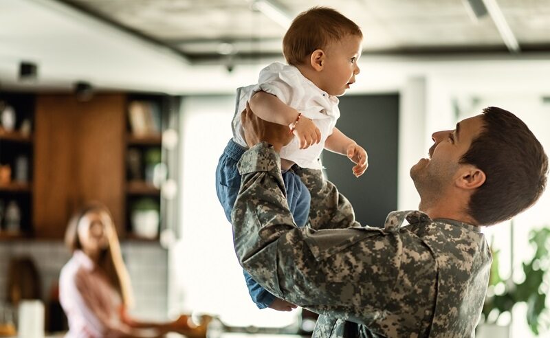 Happy army soldier playing with his baby son and having fun at home. Mother is in the background.