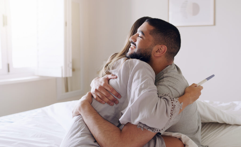 A married couple hugs while the woman holds a pregnancy test. The man is smiling and excited