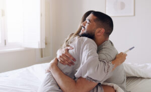A married couple hugs while the woman holds a pregnancy test. The man is smiling and excited